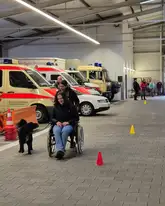 Hund läuft in einer Fahrzeughalle mit rettungswägen neben einer Frau im Rollstuhl 