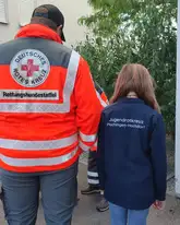 Eine Einsatzkraft der Rettungshundestaffel und ein Kind des Jugenrotkreuz von hinten
