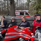 Einsatzkräfte sitzen auf einer Parkbank zugedeckt mit einer Einsatzjacke vor einem Einsatzfahrzeug